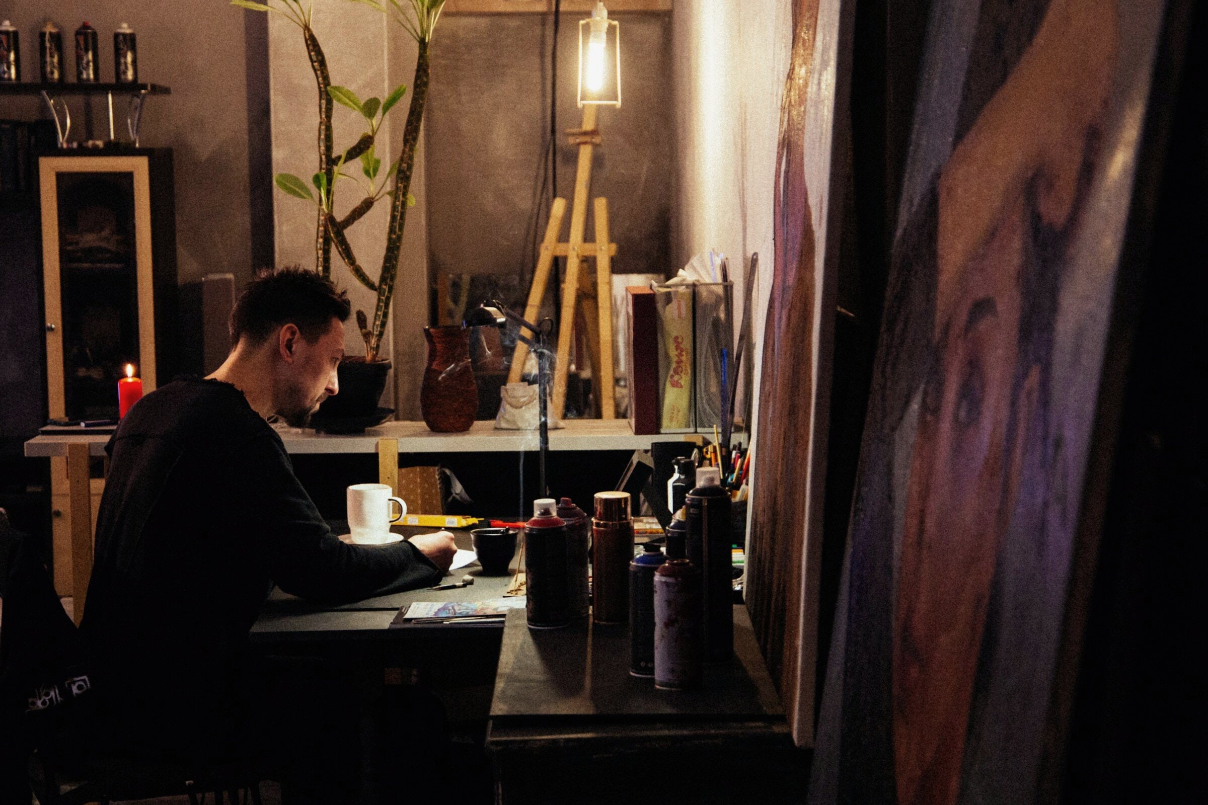 An artist drawing at a desk under a single lamp in a dark studio.