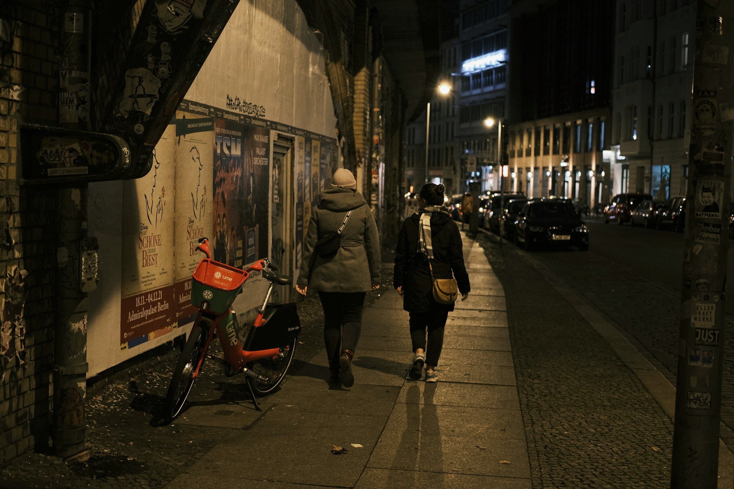 Two people walking past a poster wall on a Berlin street at night.